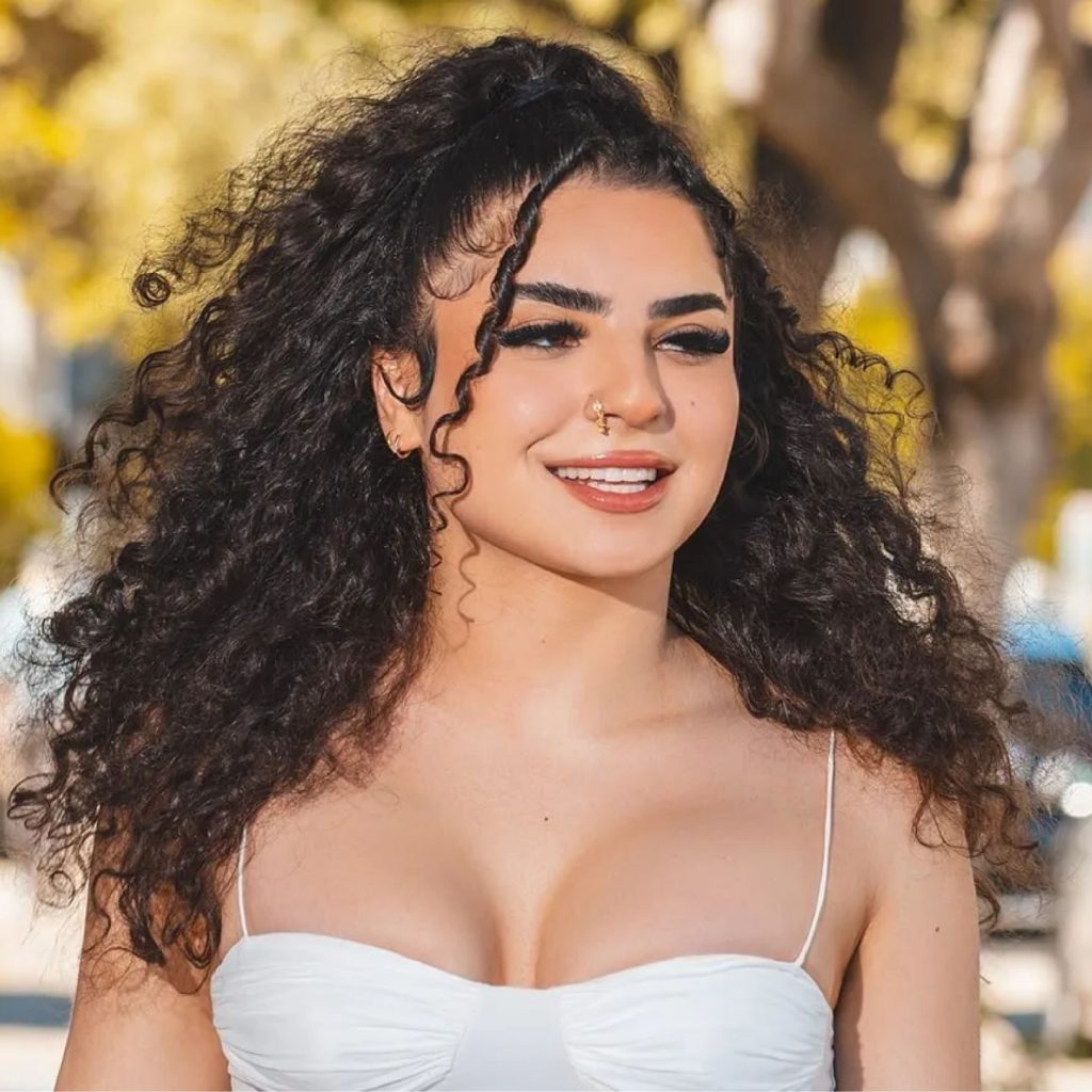 Smiling woman with curly dark hair wearing a white top outdoors.